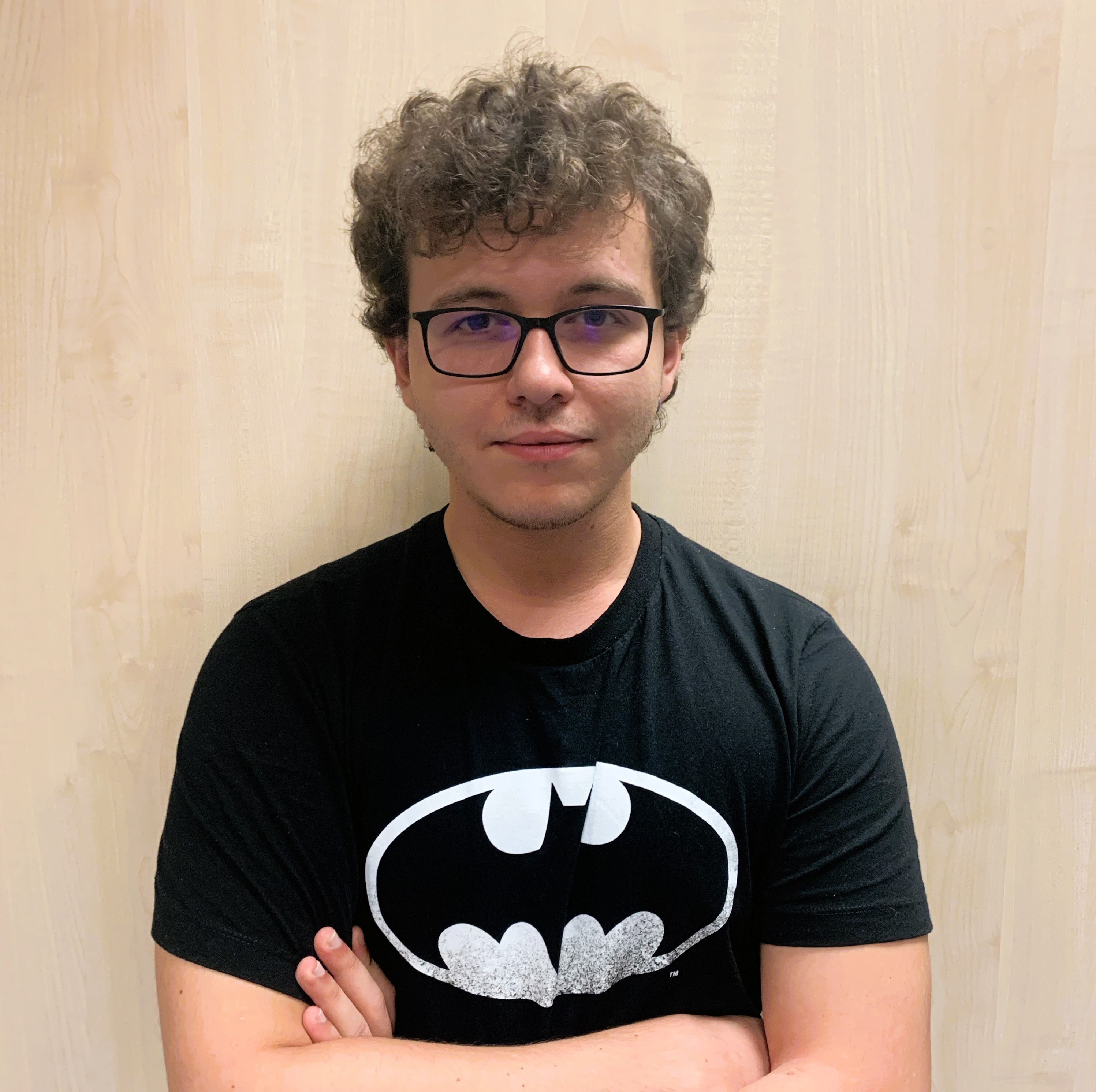 Man with curly hair and glasses wearing a black Batman T-shirt, standing with arms crossed against a light wooden background.